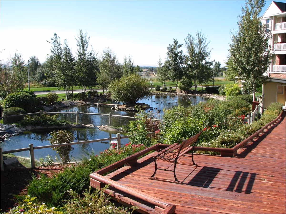 A peaceful outdoor garden area with a wooden deck featuring a bench, surrounded by lush greenery and flowering plants. There is a pond with a small island and several trees in the background under a clear blue sky.