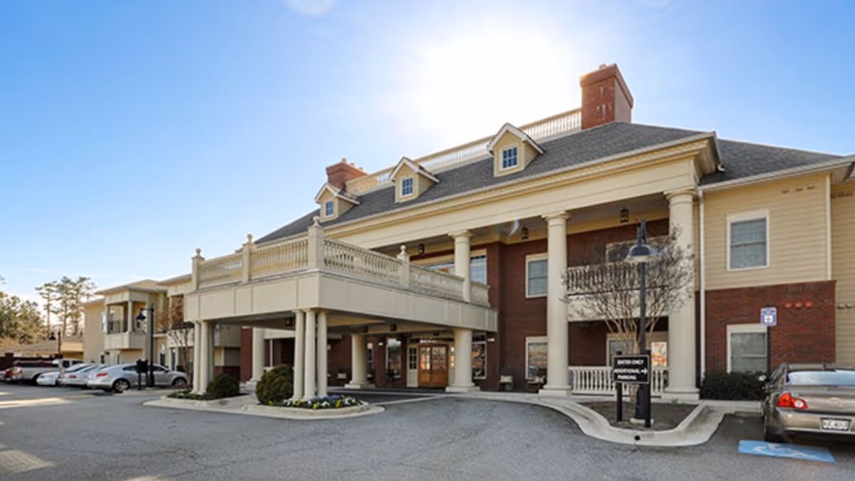 Exterior view of a senior living facility building with a covered entrance supported by large white columns, multiple windows, and a parking area with several cars parked. The building has a combination of brick and light-colored siding with a clear blue sky in the background.