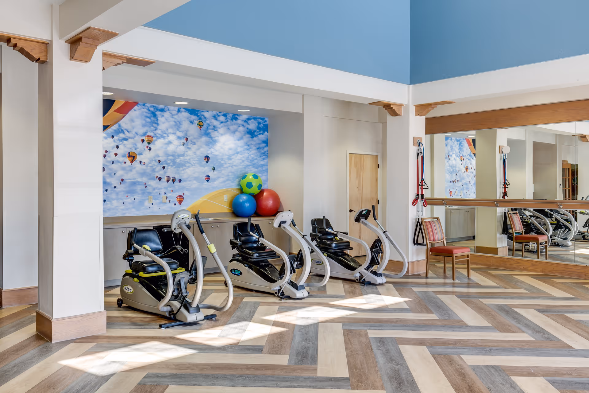 A bright exercise room with three recumbent exercise bikes positioned on a patterned wooden floor. The back wall features a colorful mural of hot air balloons in a blue sky with clouds. There are large mirrors on the right wall, a wooden chair, and some exercise equipment including resistance bands hanging on the wall.