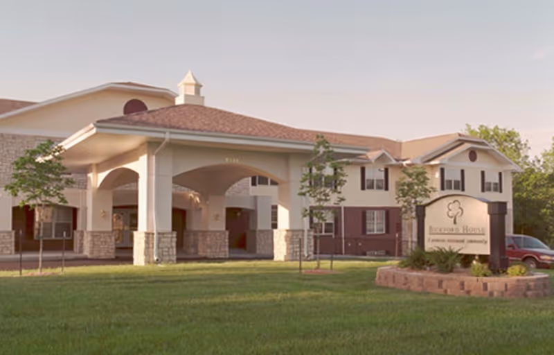 Exterior view of a senior living facility building with a covered entrance, stone and beige siding, and a sign in front reading 'Beechford House, a private retirement community'. The building is surrounded by a well-maintained lawn and small trees.