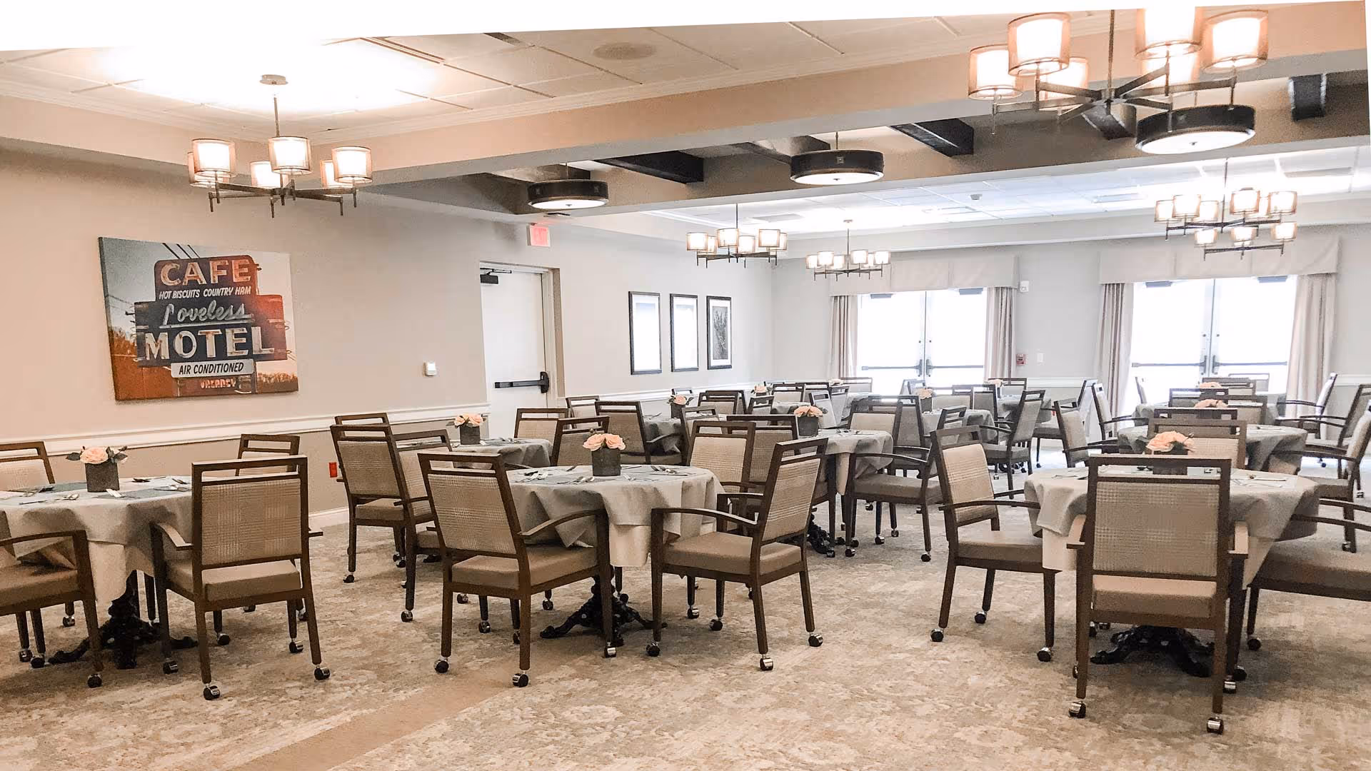 A spacious dining room with multiple round tables covered with light-colored tablecloths, each adorned with a small flower arrangement. The room features beige walls, large windows with curtains, and modern ceiling light fixtures. There is a vintage-style wall art piece on the left wall that reads 'CAFE Loveless MOTEL'.