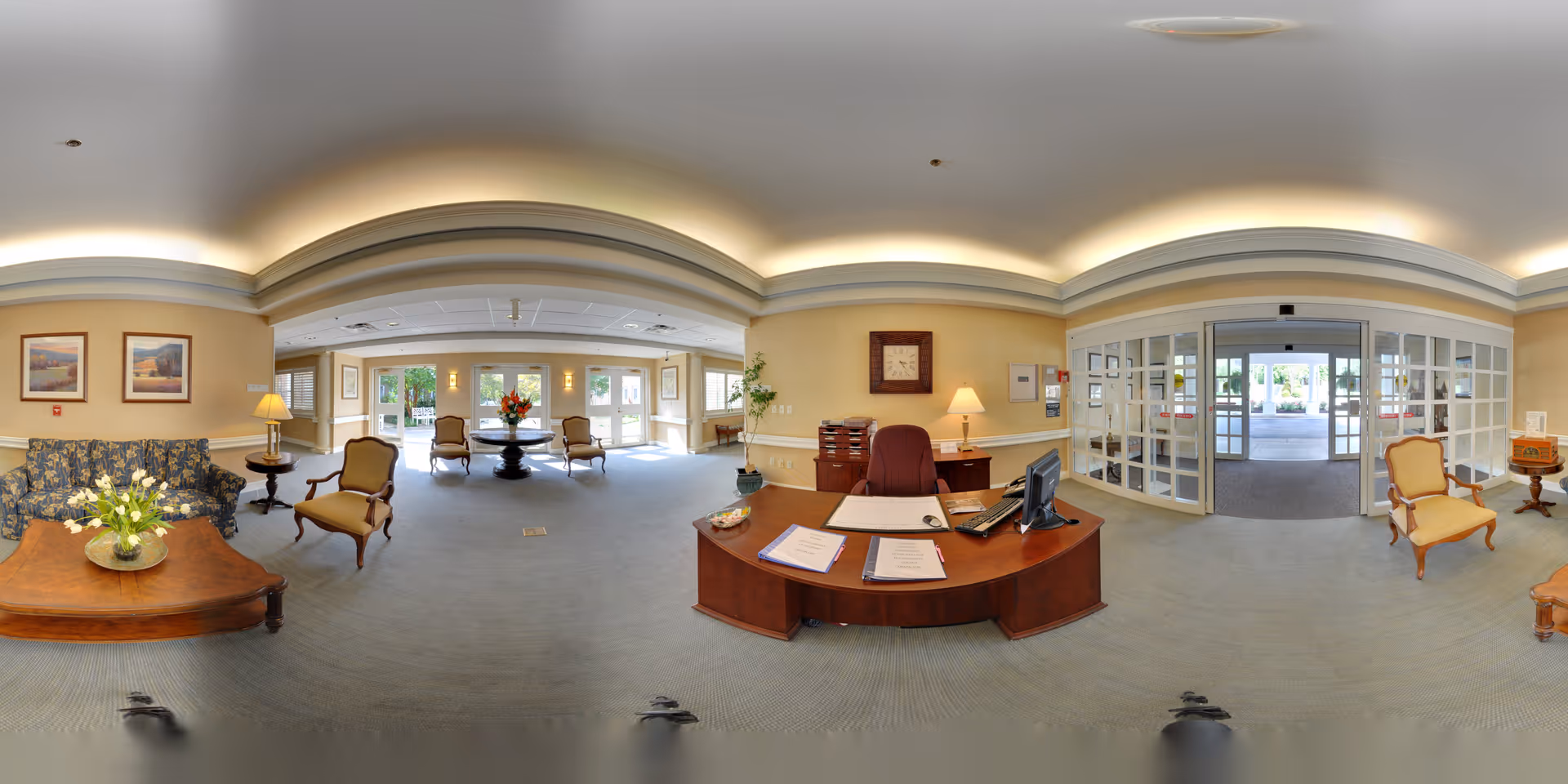 Spacious, well-lit reception lobby with a central wooden front desk, seating areas, floral arrangements and glass entrance doors.