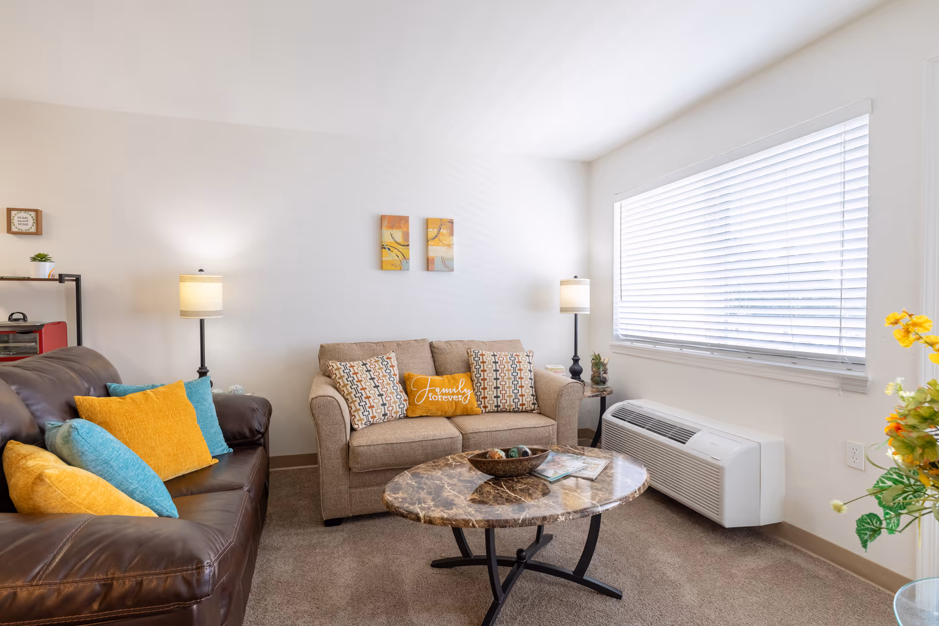 A cozy living room with a brown leather sofa and a beige fabric loveseat, both adorned with colorful throw pillows in yellow, blue, and patterned designs. A round marble-top coffee table sits in the center with decorative items on it. Two floor lamps with beige shades flank the loveseat, and a large window with white blinds lets in natural light. The walls are white with two small abstract paintings above the loveseat. A small shelf with a plant and decorative items is visible on the left side, and a heating/cooling unit is beneath the window on the right.