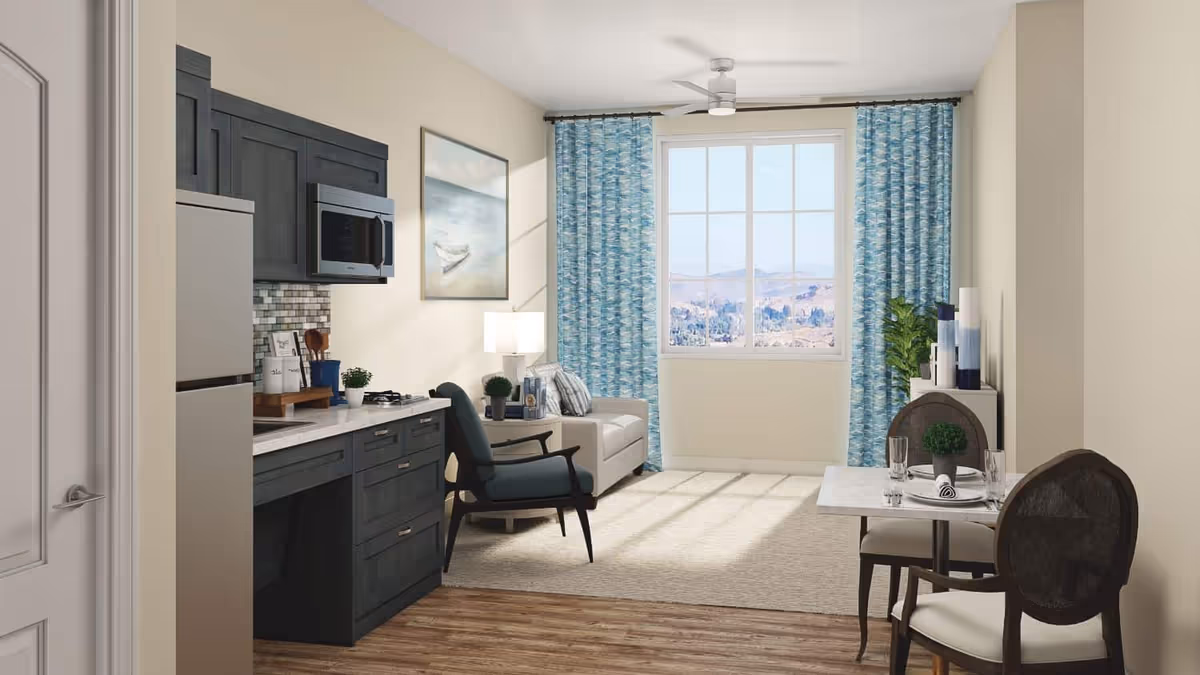 Bright studio living area with a kitchenette on the left, seating and a small dining table by a large window with blue curtains.