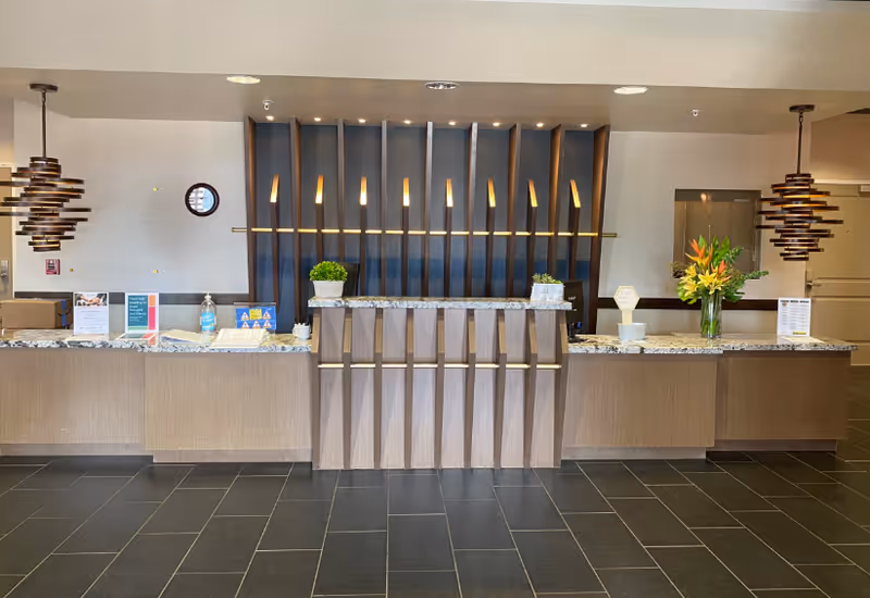 Reception desk area in a senior living facility with a modern design. The desk has a granite countertop with two potted plants and a vase of flowers. Behind the desk is a decorative wooden panel with vertical slats and warm lighting. Two pendant lights hang from the ceiling on either side of the desk. The floor is tiled in dark gray.