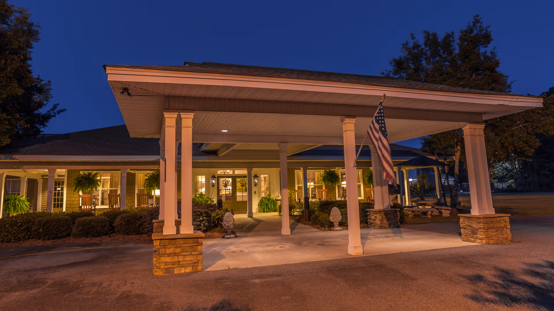 Exterior view of Fellowship Nashville Assisted Living Community at dusk, showing the covered entrance with pillars, an American flag, and warm lights glowing from inside the building surrounded by greenery.