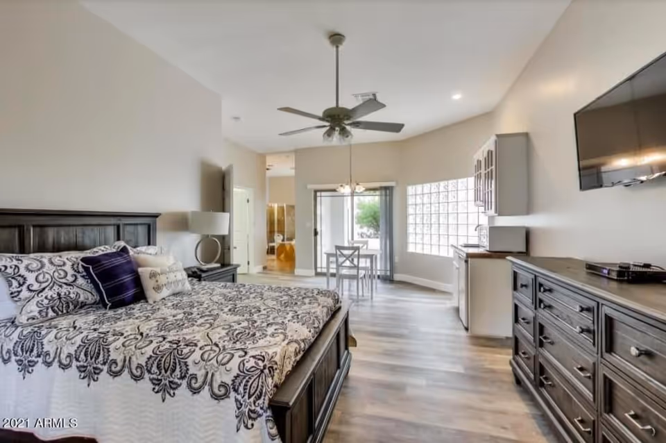 A spacious bedroom with a large bed featuring patterned bedding and multiple pillows. The room includes a dark wooden dresser with a flat-screen TV mounted above it, a nightstand with a lamp, and a small dining table with chairs near a sliding glass door leading outside. The room has light-colored walls, wood flooring, a ceiling fan, and a kitchenette area with cabinets and a microwave.