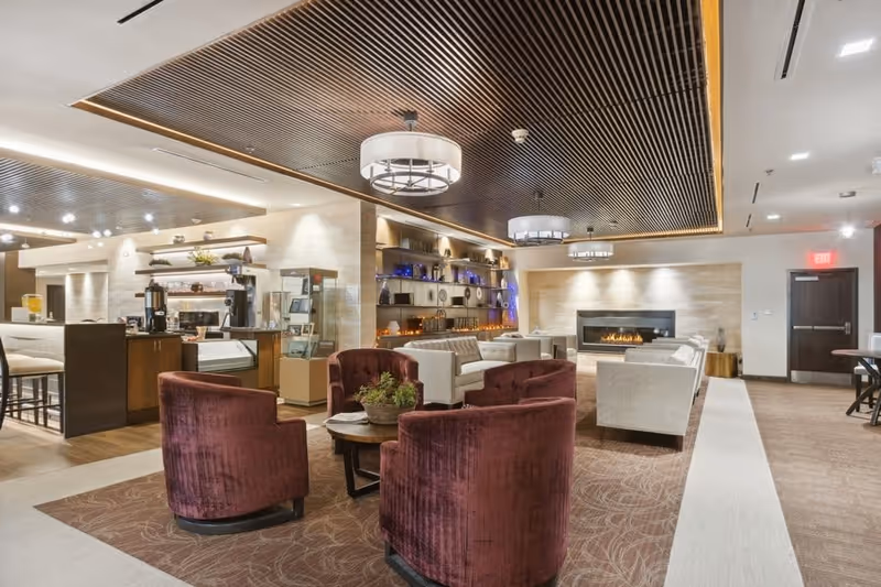 A modern senior living facility common area with plush burgundy armchairs and beige sofas arranged around a coffee table. The room features a decorative fireplace, built-in shelves with various ornaments, and a ceiling with wooden slats and circular light fixtures. There is a small kitchenette or coffee station area to the left with bar stools and a counter.