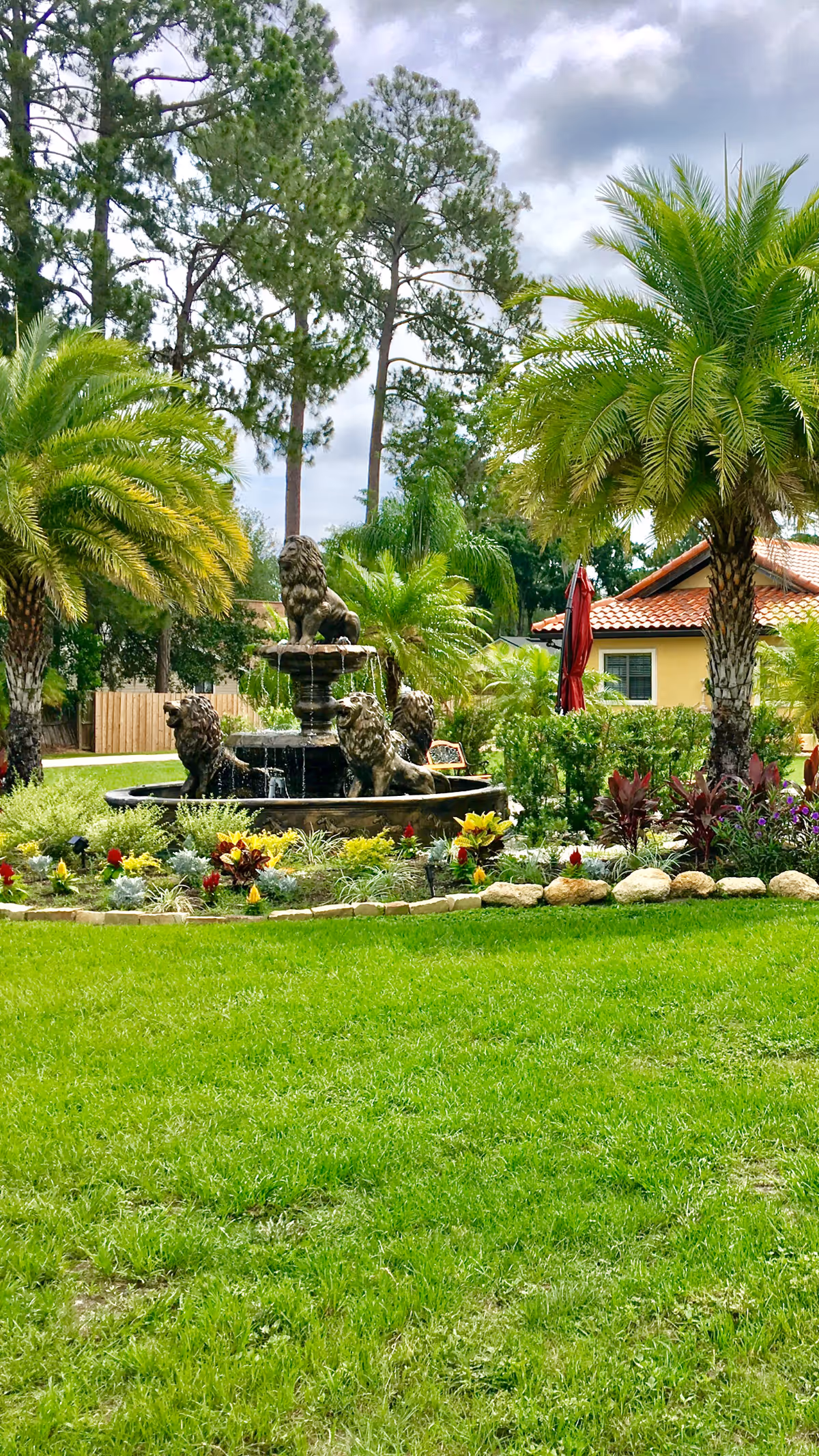 A lush green garden area with a decorative fountain featuring lion statues in the center. Surrounding the fountain are various colorful flowers and plants. Tall palm trees and pine trees are visible in the background, along with a yellow building with a red-tiled roof and a red umbrella.