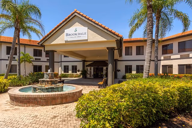 Exterior view of Brookdale Phillippi Creek senior living facility with a covered entrance, a circular water fountain in front, palm trees, and well-maintained landscaping under a clear blue sky.