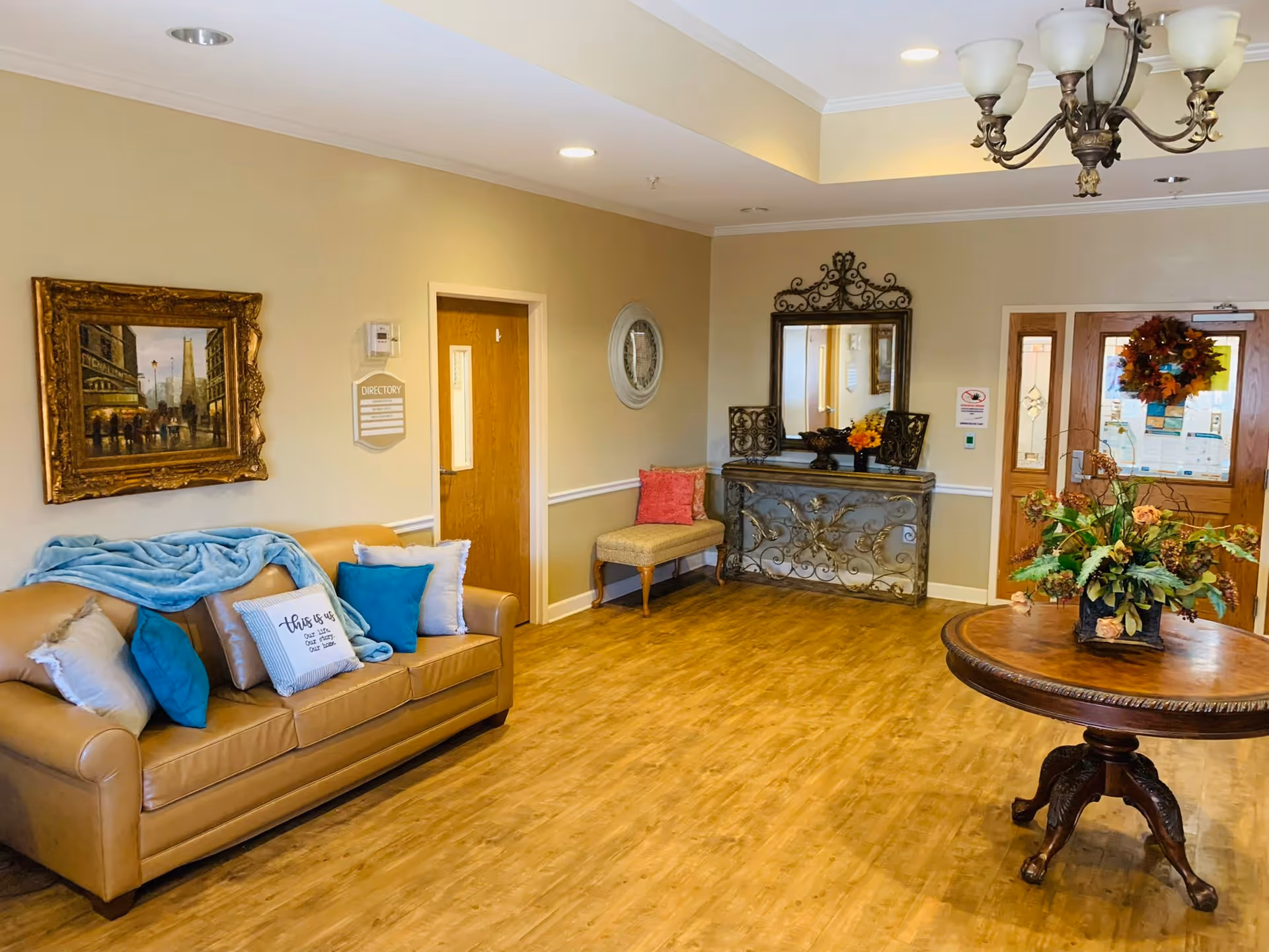 A cozy and well-lit interior common area with a tan leather sofa adorned with blue and white pillows and a blue throw blanket. There is a framed painting on the wall above the sofa, a wooden door with a directory sign next to it, a decorative console table with a large ornate mirror above it, and a small bench with a red pillow. A round wooden table with a floral arrangement is in the foreground, and double wooden doors with glass panels and a wreath are visible in the background.