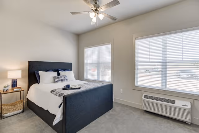 A bright and clean bedroom with a dark upholstered bed frame, white bedding, and decorative pillows. There is a small side table with a lamp, books, and a plant. Two large windows with white blinds let in natural light. A ceiling fan with lights is mounted on the ceiling, and a wall-mounted air conditioning unit is below one window.