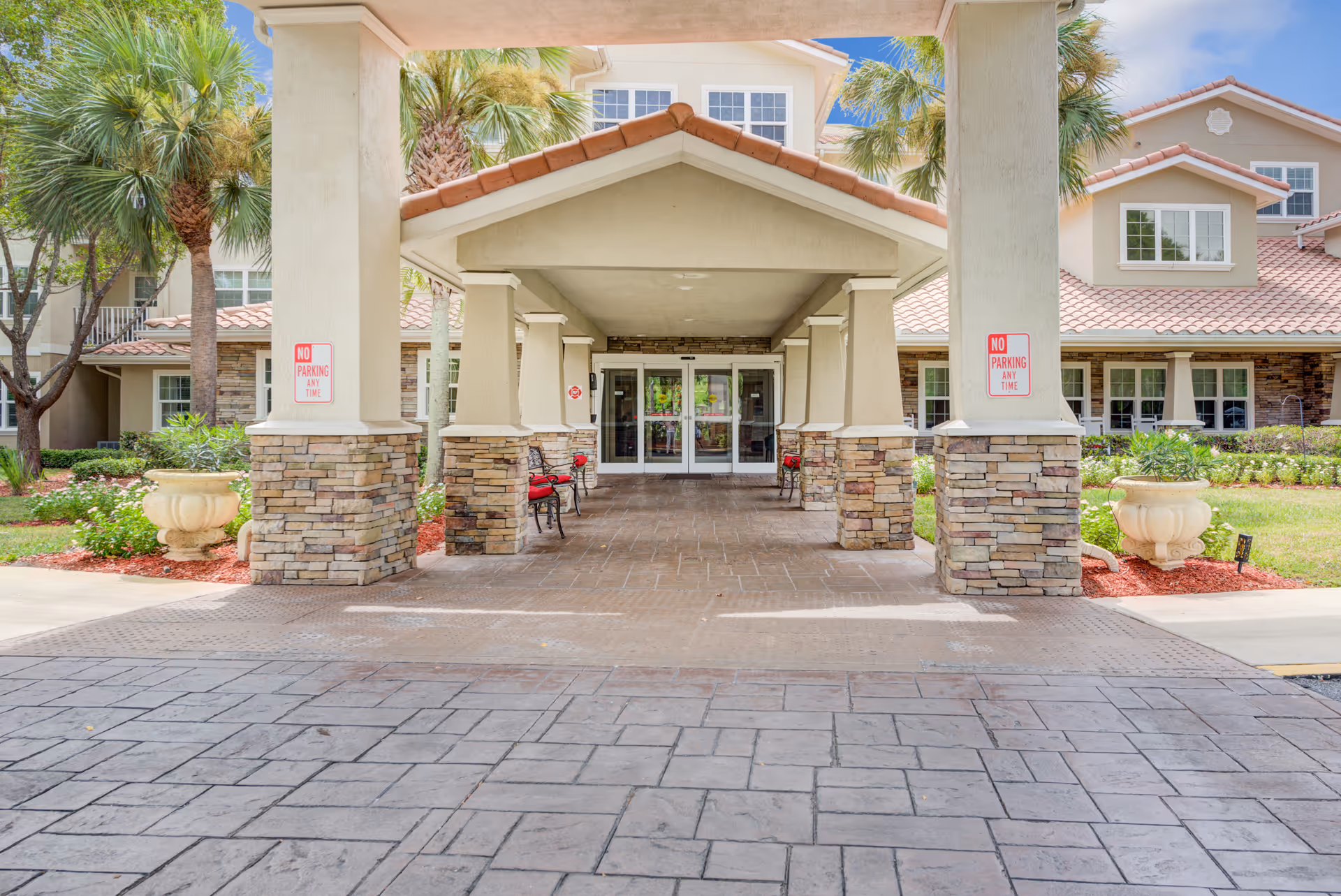 Entrance to a senior living facility with a covered driveway supported by stone and stucco columns. There are red cushioned benches along the walkway, palm trees and landscaping on either side, and a building with multiple windows and a tiled roof in the background.