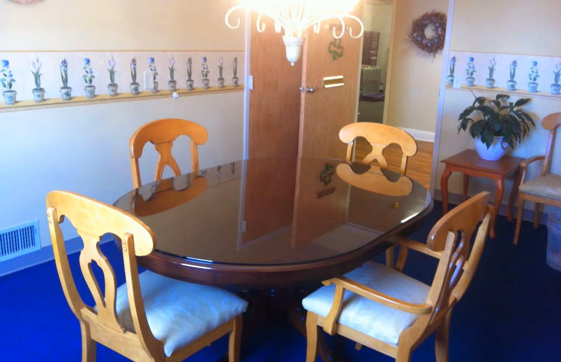 A dining room with a dark wooden oval table surrounded by four wooden chairs with light-colored cushions. The room has blue carpet, light-colored walls with a floral wallpaper border, a wooden door, a small wooden side table with a potted plant, and a decorative wreath on the wall.