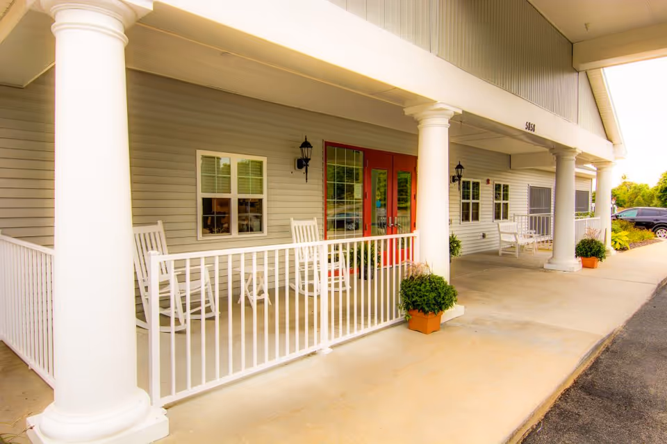 Front porch area of a building with white columns, white railing, rocking chairs, potted plants, and red double doors under a covered walkway.