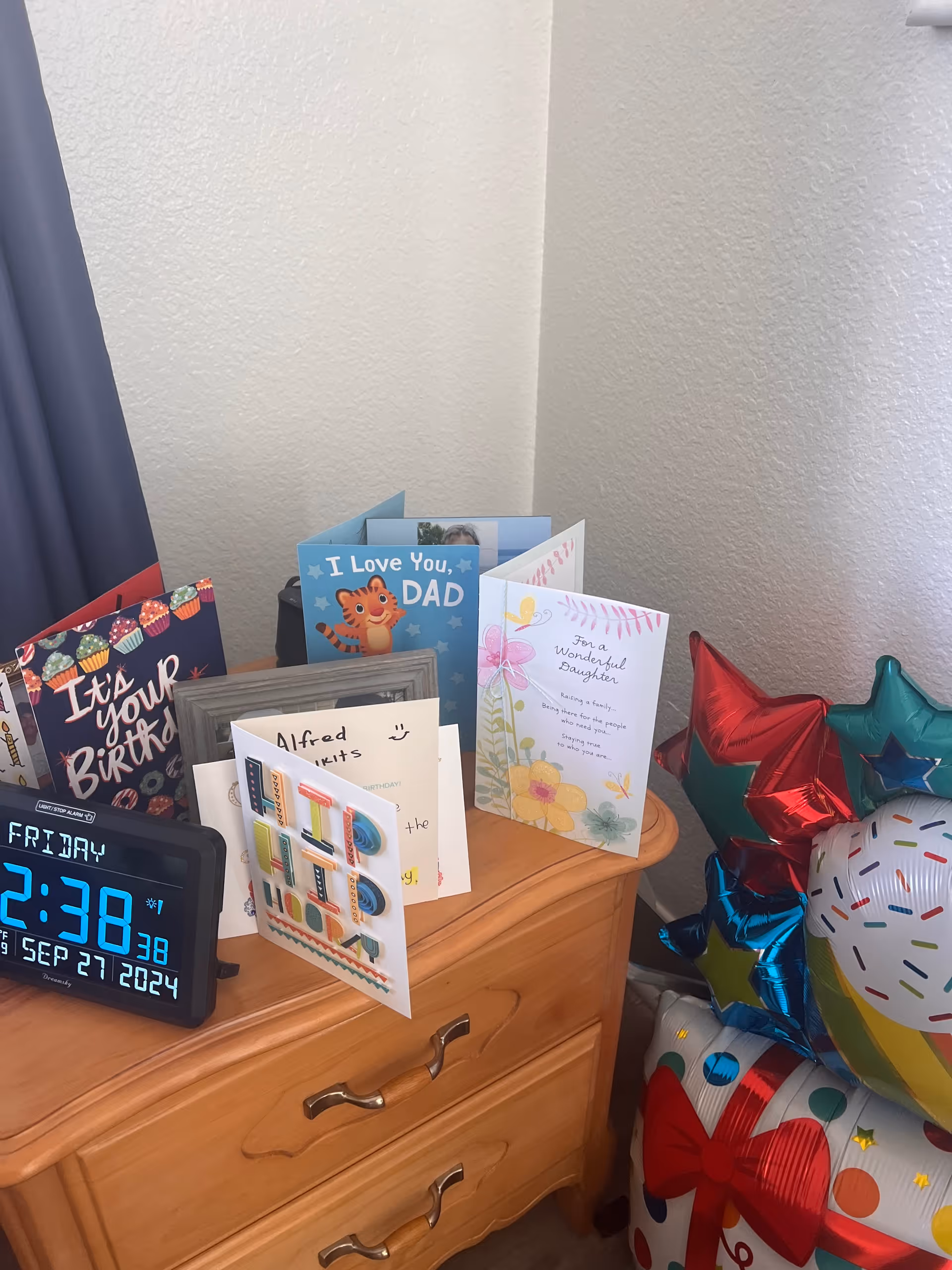 A wooden nightstand with several greeting cards displayed on top, including birthday and Father's Day cards. A digital clock showing the date and time is also on the nightstand. To the right, there are colorful star-shaped and birthday-themed balloons. The background shows a plain white textured wall and a dark curtain on the left side.