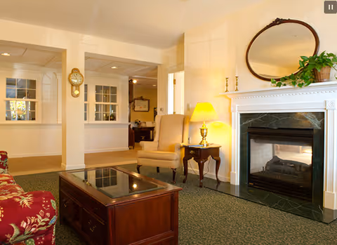 A cozy living room area featuring a floral patterned sofa, a wooden coffee table with a glass top, an armchair next to a side table with a lamp, and a fireplace with a black marble surround and a white mantel. Above the mantel is an oval mirror and a potted plant. The room has beige walls and carpeted flooring, with a grandfather clock visible in the background.