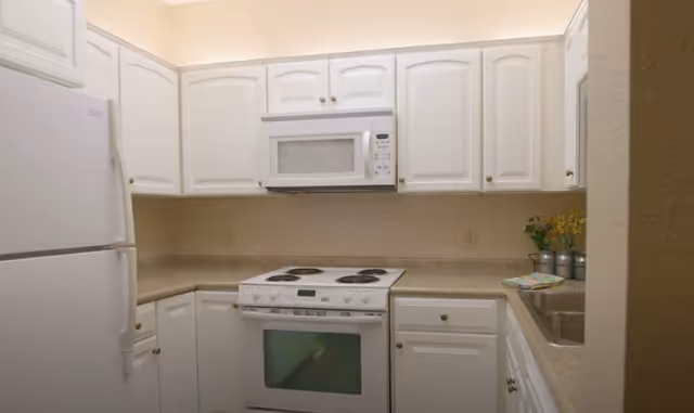 A compact kitchen with white cabinets, a white refrigerator, a white electric stove with oven, a white microwave mounted above the stove, and a countertop with a sink. There are small decorative plants and a folded cloth on the counter near the sink.