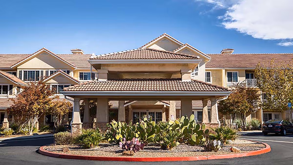 Front exterior view of Holiday Rancho Village senior living facility with a covered entrance, desert landscaping including cacti, and a clear blue sky.