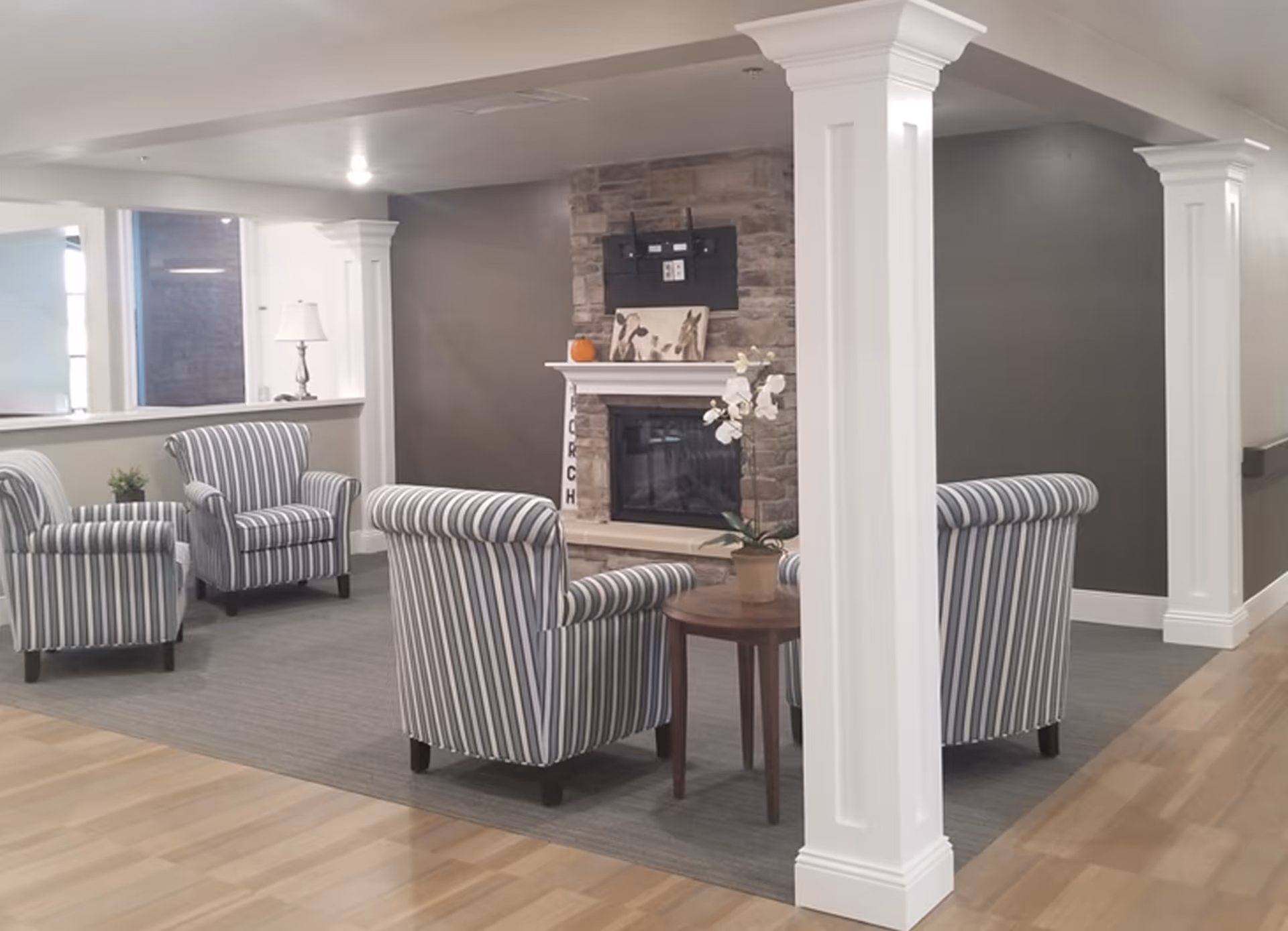 A cozy sitting area in a senior living facility featuring four striped armchairs arranged around a small round wooden table with a potted orchid. The space includes a stone fireplace with a TV mount above it, a small pumpkin decoration, and a lamp on a side table near a window. The room has light wood flooring, a gray carpeted area, and white pillars.