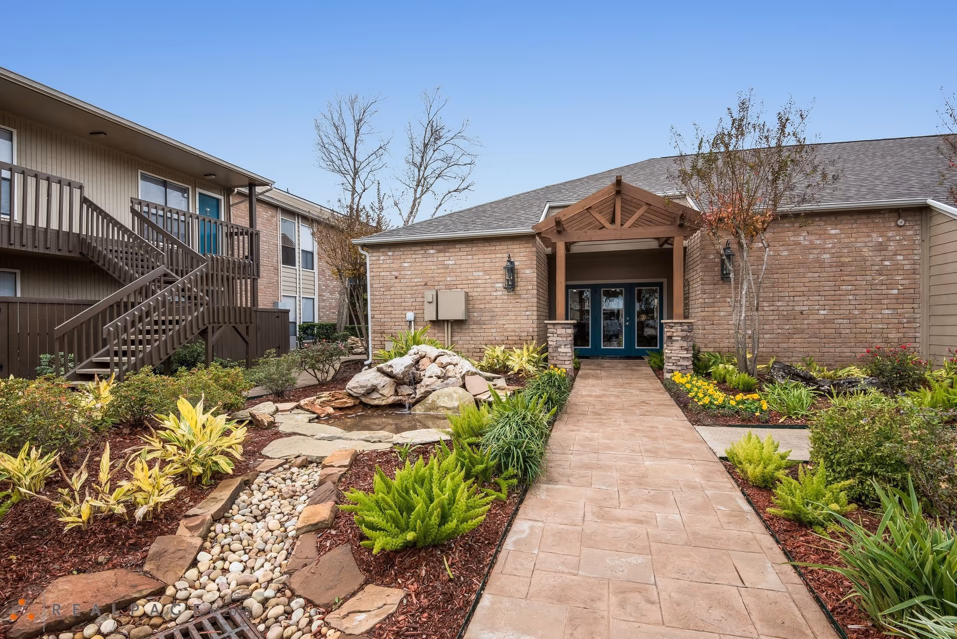 Exterior view of Braesridge Apartments showing a paved walkway leading to a building entrance with double glass doors under a wooden pergola. The surrounding landscaping includes various green plants, bushes, a small rock water feature, and a tree with some autumn leaves. To the left, there is a two-story building with brown stairs and railings.