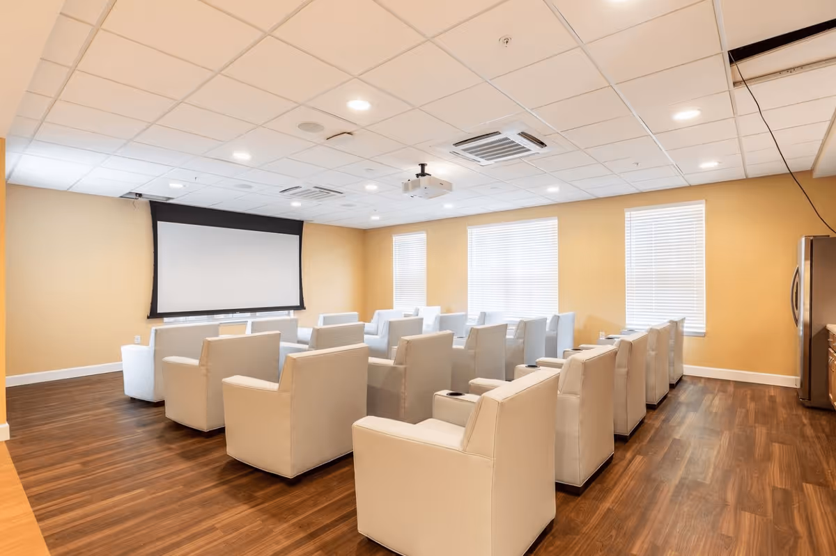 A room with multiple rows of beige armchairs facing a large projector screen mounted on a yellow wall. The room has wooden flooring, three windows with blinds, a ceiling-mounted projector, and recessed lighting in the ceiling.