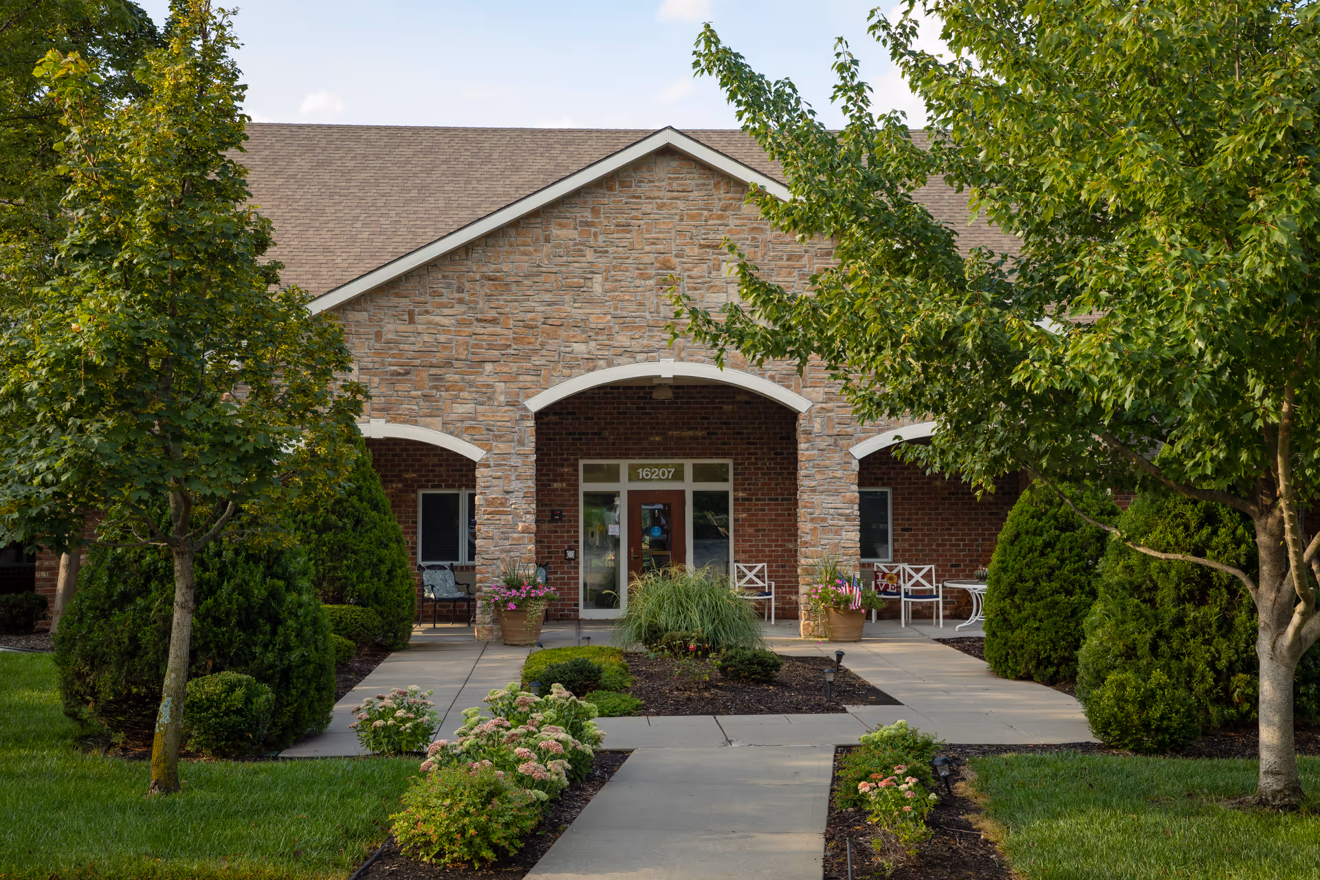 Front entrance of a brick-and-stone assisted living building with a landscaped walkway, arched porch, and outdoor seating.