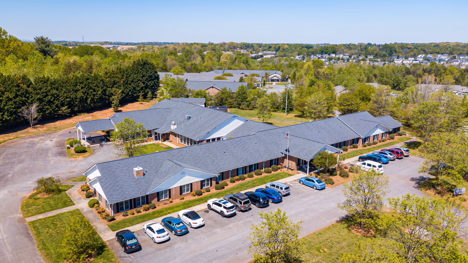 Aerial view of a senior living facility with multiple connected single-story buildings featuring gray roofs and brick walls. Several cars are parked in front of the buildings, surrounded by green trees and landscaped areas under a clear blue sky.