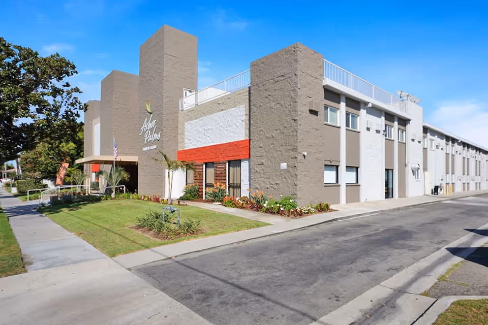 Exterior view of a modern two-story building with beige and white walls, a red accent stripe, and a well-maintained lawn with flowers and small palm trees. The building has a sign that reads 'Arbor Palms' and an American flag near the entrance. The sky is clear and blue.