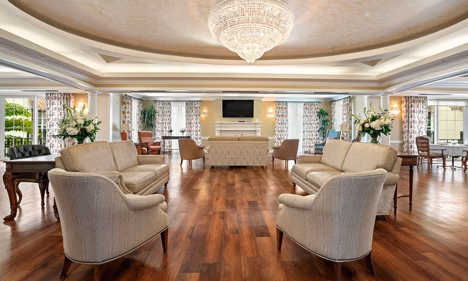 Spacious and elegant living room area with wooden flooring, beige sofas and armchairs arranged around a central fireplace with a mounted TV above it. Large windows with floral curtains allow natural light to fill the room, and a grand chandelier hangs from the ceiling. Side tables with flower arrangements and additional seating are visible in the background.