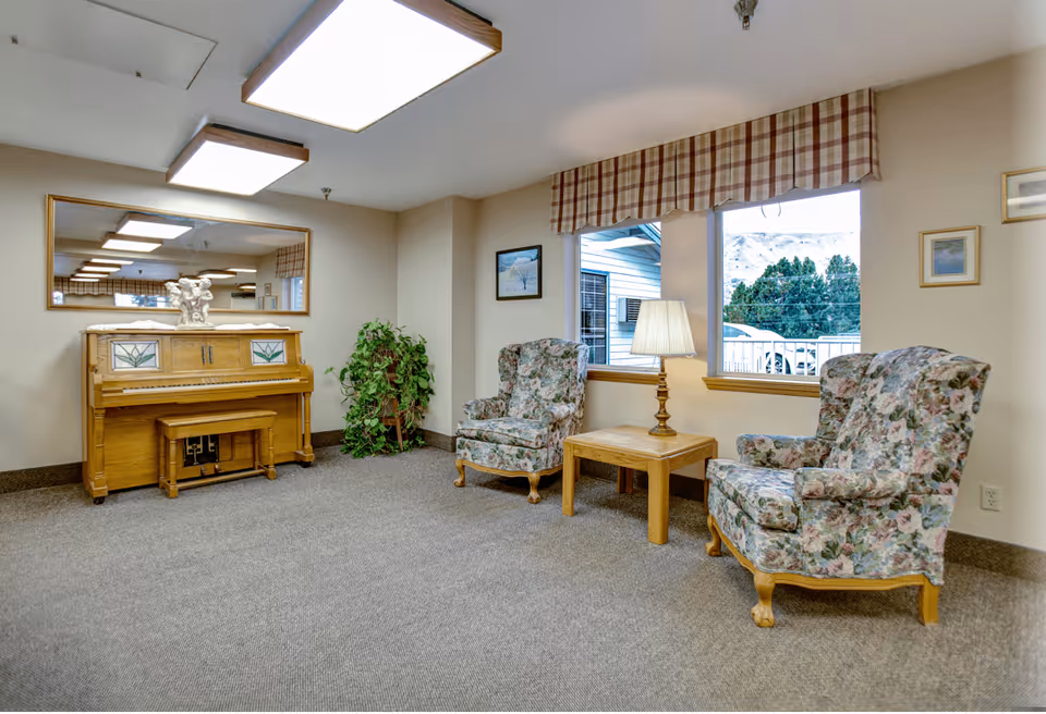 A cozy sitting area in a senior living facility featuring two floral upholstered armchairs with wooden legs, a wooden side table with a lamp, a wooden piano with a matching bench, a large wall mirror above the piano, a potted plant, and two windows with plaid valances showing an outdoor view.