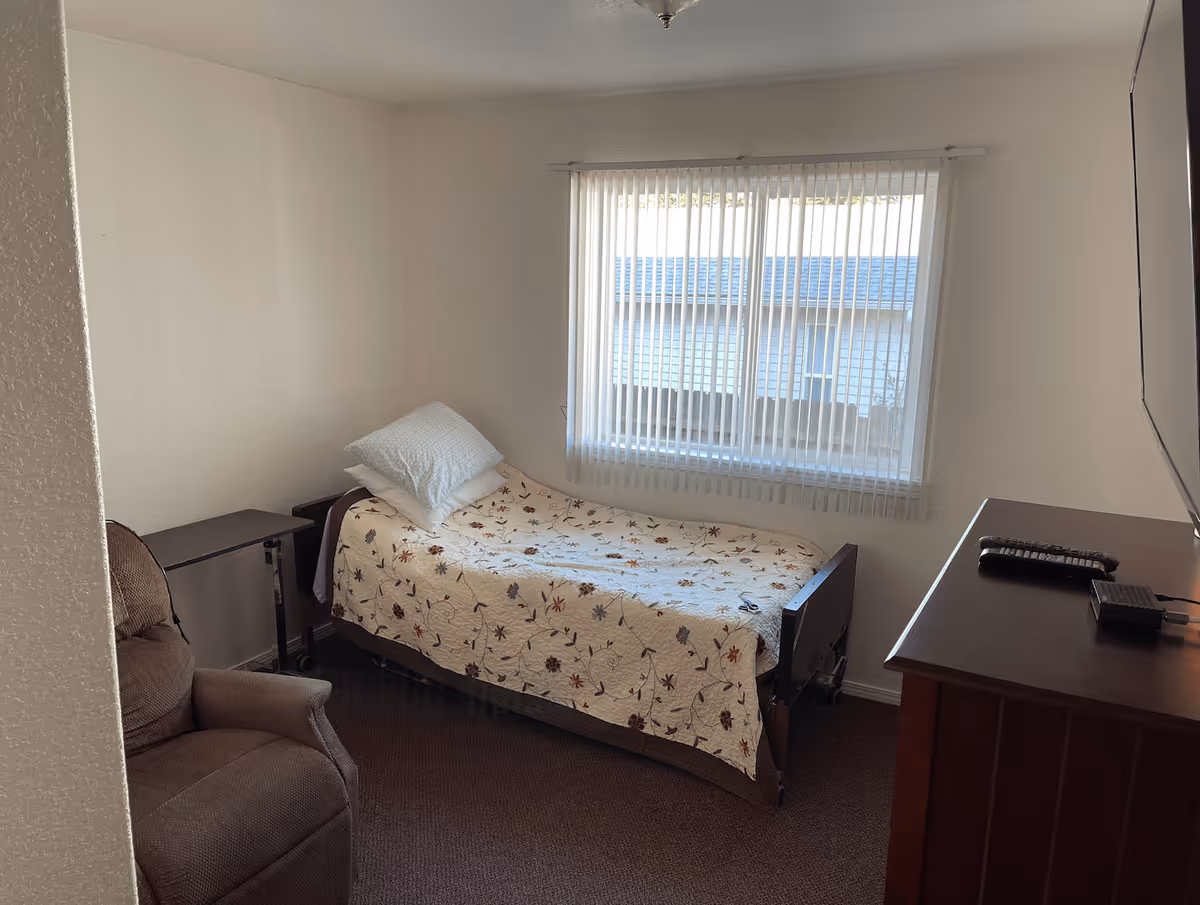 A small bedroom with a single bed covered with a floral quilt, two pillows, a brown recliner chair, a small adjustable table, a wooden dresser with a TV on top, and a window with vertical blinds letting in natural light.