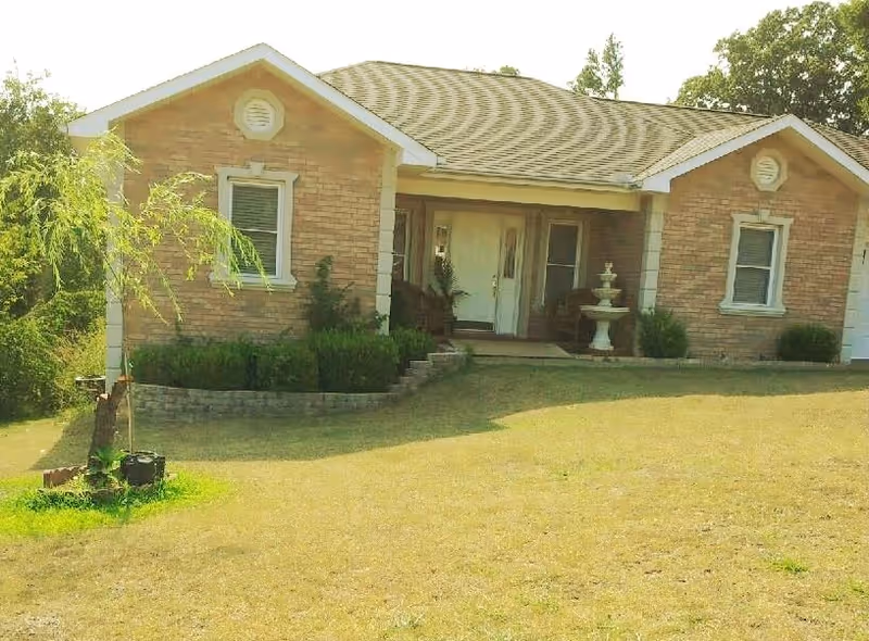 Single-story brick house with a gabled roof, two windows on either side of a central front door, a small porch with chairs, a white decorative fountain, and a well-maintained lawn with some shrubs and a small tree in front.