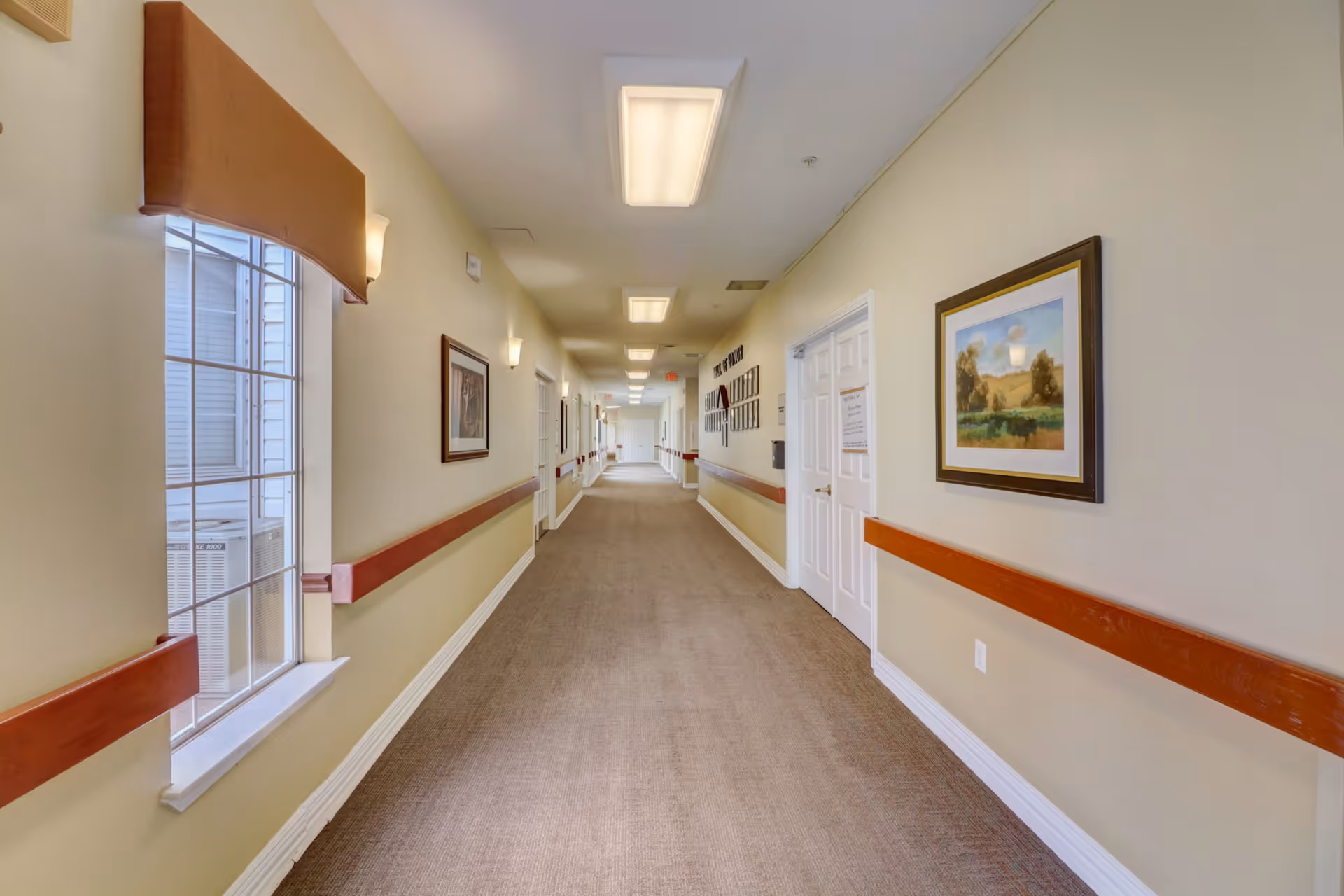 Long indoor hallway in a senior living facility with beige walls, carpeted floor, wooden handrails on both sides, framed artwork on the walls, and ceiling lights. A large window on the left side lets in natural light.