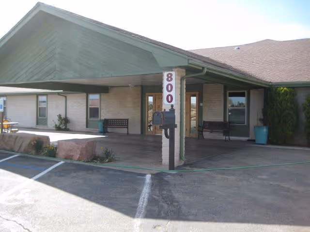 Exterior view of a single-story building with a covered entrance supported by a pillar displaying the number 800. There are benches near the entrance, a mailbox attached to the pillar, and some potted plants. The area in front includes a parking lot with marked spaces.