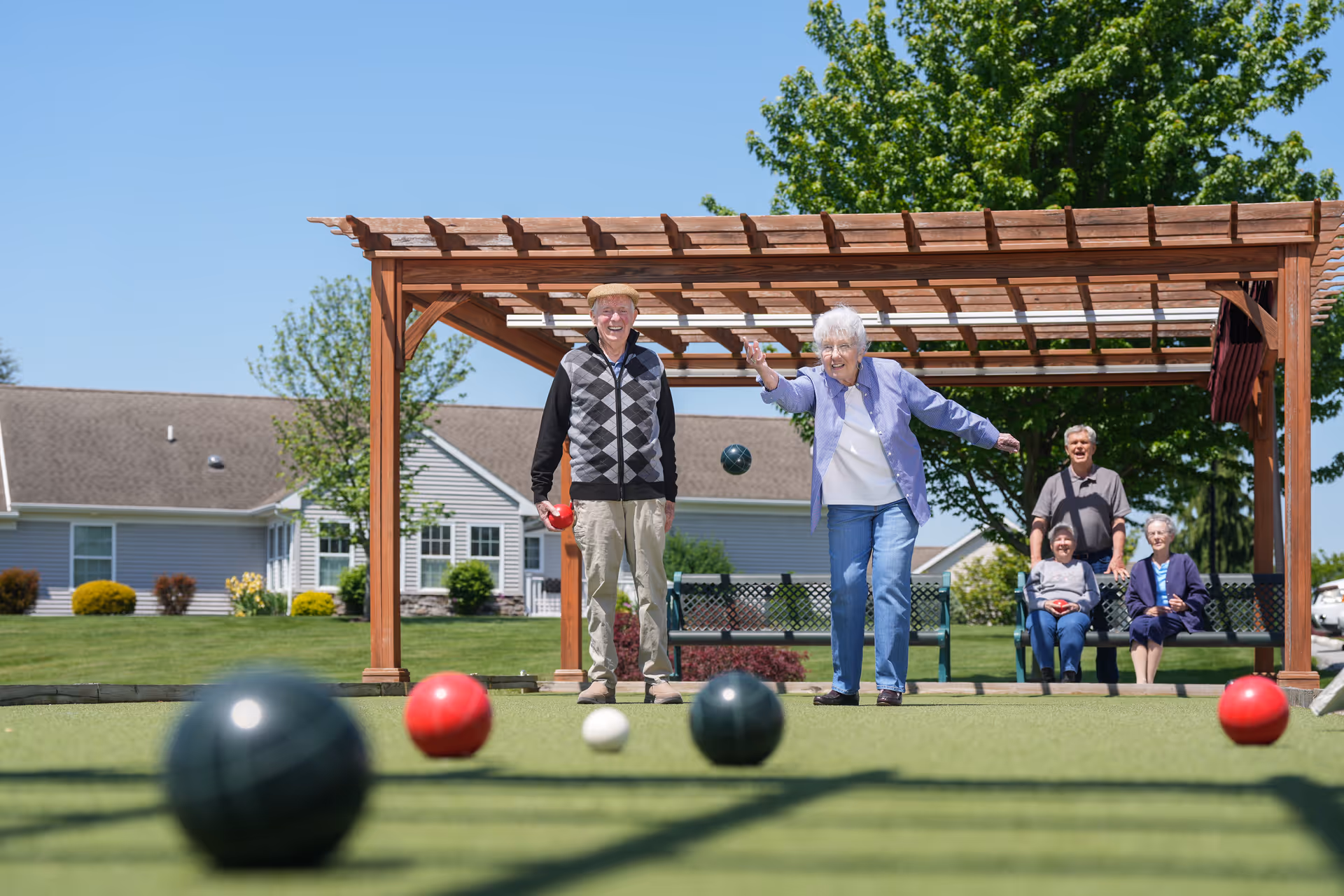 Two elderly people playing bocce ball on a green lawn under a wooden pergola, with three other elderly people sitting on benches in the background, and residential buildings and trees visible on a sunny day.