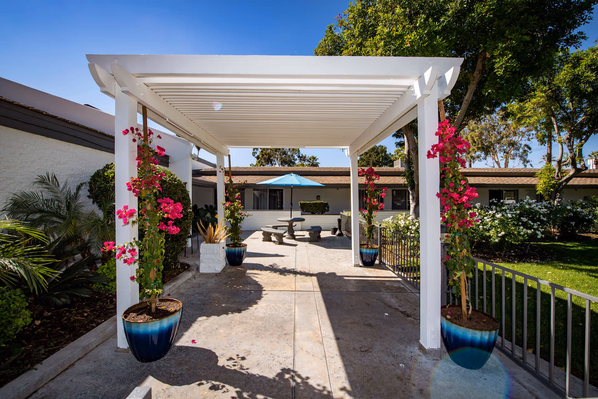 Outdoor patio area at Community Care & Rehabilitation Center featuring a white pergola with pink flowering plants in blue pots climbing its columns, a concrete walkway, green bushes, trees, and a round stone table with benches under a blue umbrella in the background.