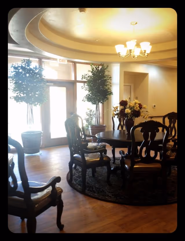 A well-lit dining area with a round wooden table surrounded by six ornate wooden chairs with cushioned seats. A floral centerpiece decorates the table. The room has wooden flooring and a circular rug under the table. Two large potted plants are placed near glass doors that allow natural light to enter. A chandelier with multiple lights hangs from the ceiling.