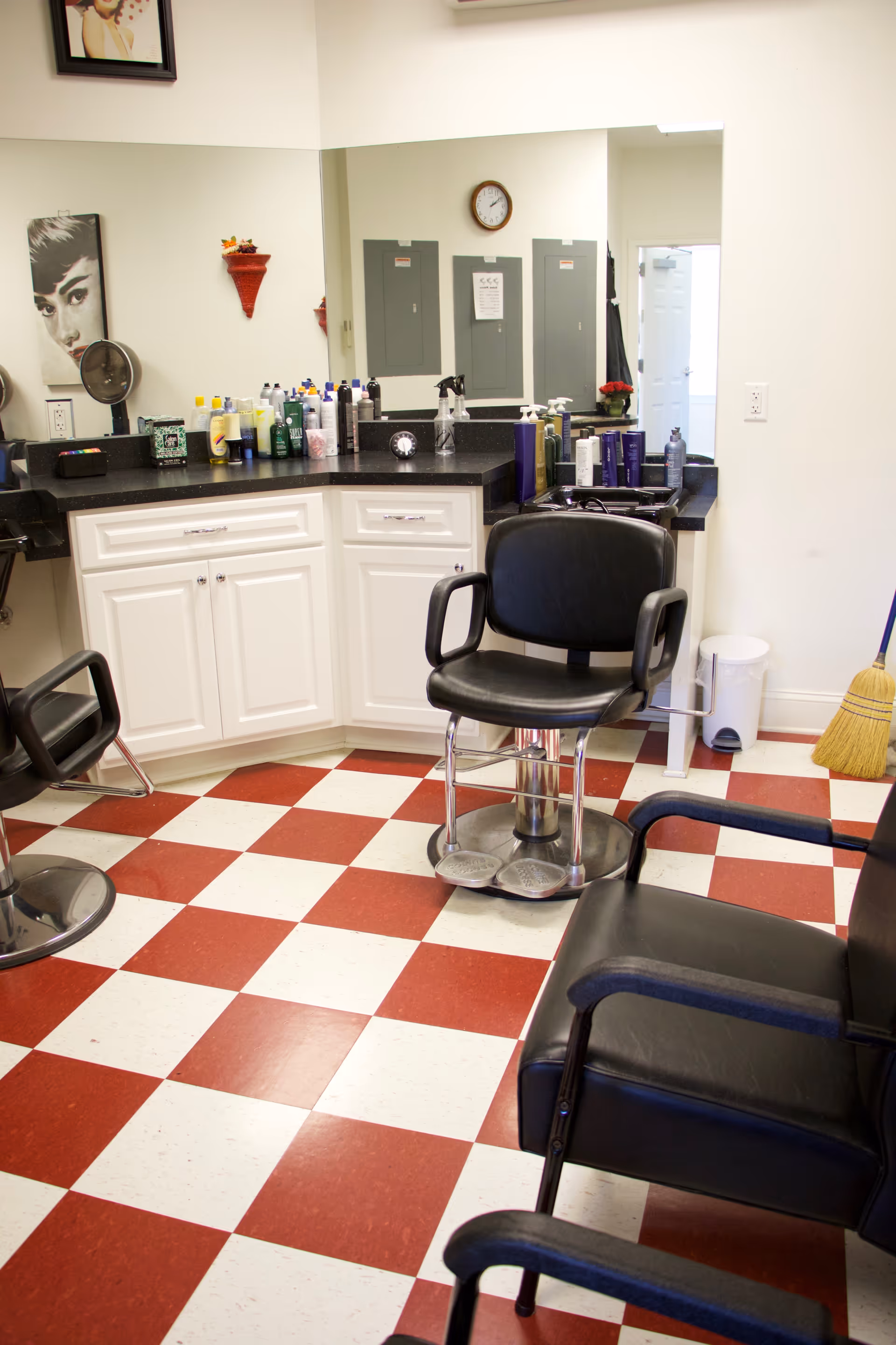 Interior salon area with barber chairs, a large mirror, countertop of hair products and a red-and-white checkerboard floor.