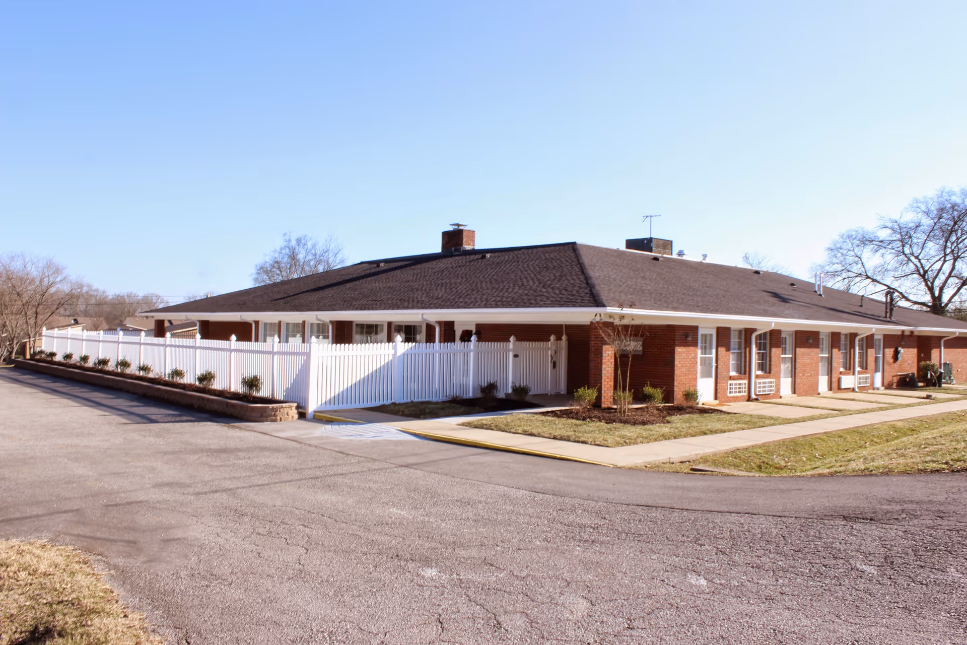 Exterior view of a single-story brick building with a dark shingled roof, white fencing, and a paved driveway, surrounded by grass and trees under a clear blue sky.