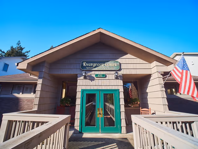 Front entrance of Evergreen Court facility with a peaked roof, green double doors, and an American flag on the right side. There is a wooden ramp leading up to the entrance and a sign above the doors that reads 'Evergreen Court'.