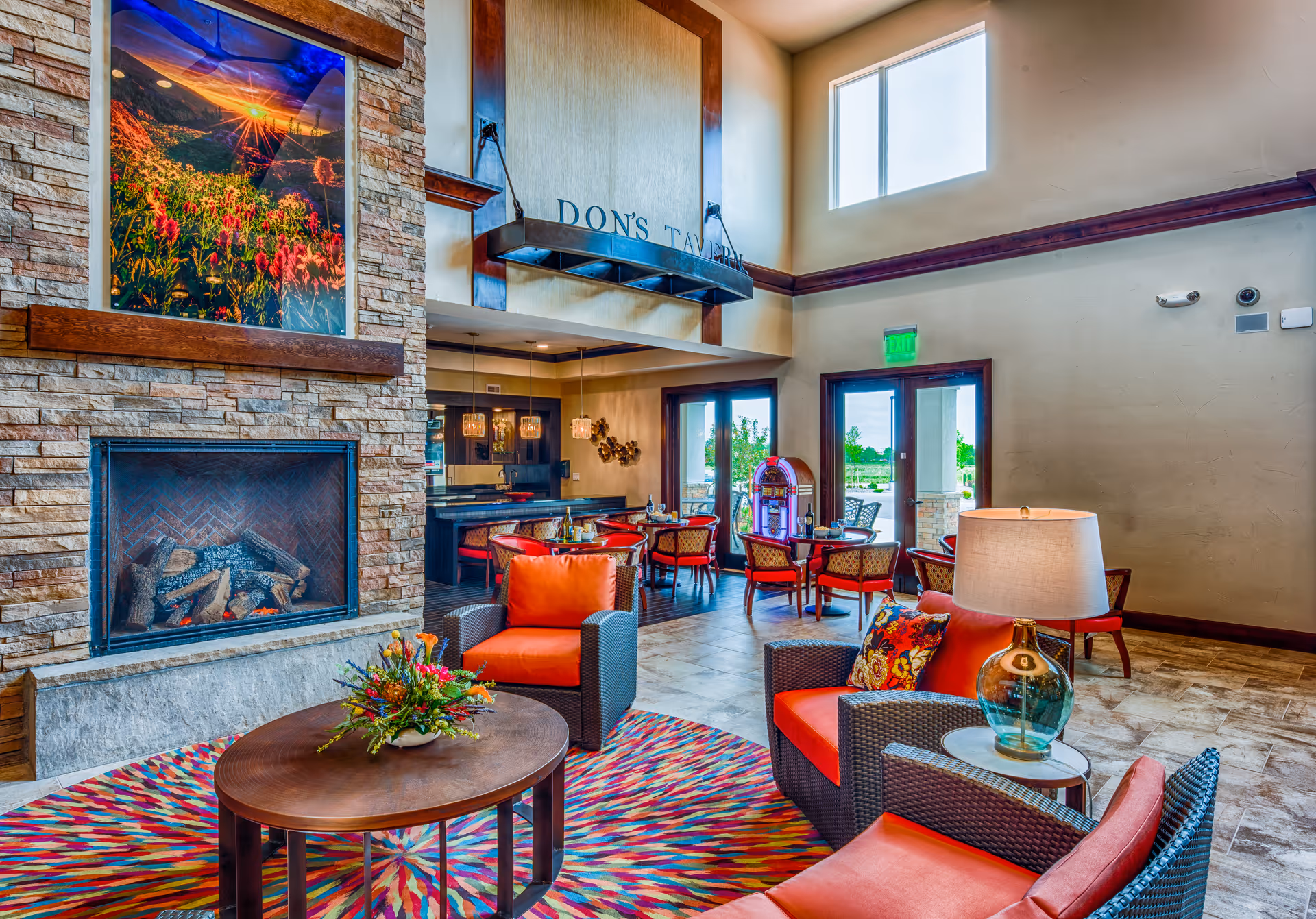 A bright and spacious assisted living common area with a stone fireplace, colorful patterned rug, and comfortable wicker chairs with orange cushions. A round wooden coffee table with a floral arrangement sits in the center. In the background, there are dining tables and chairs near large windows and glass doors letting in natural light. A jukebox is visible near the dining area.