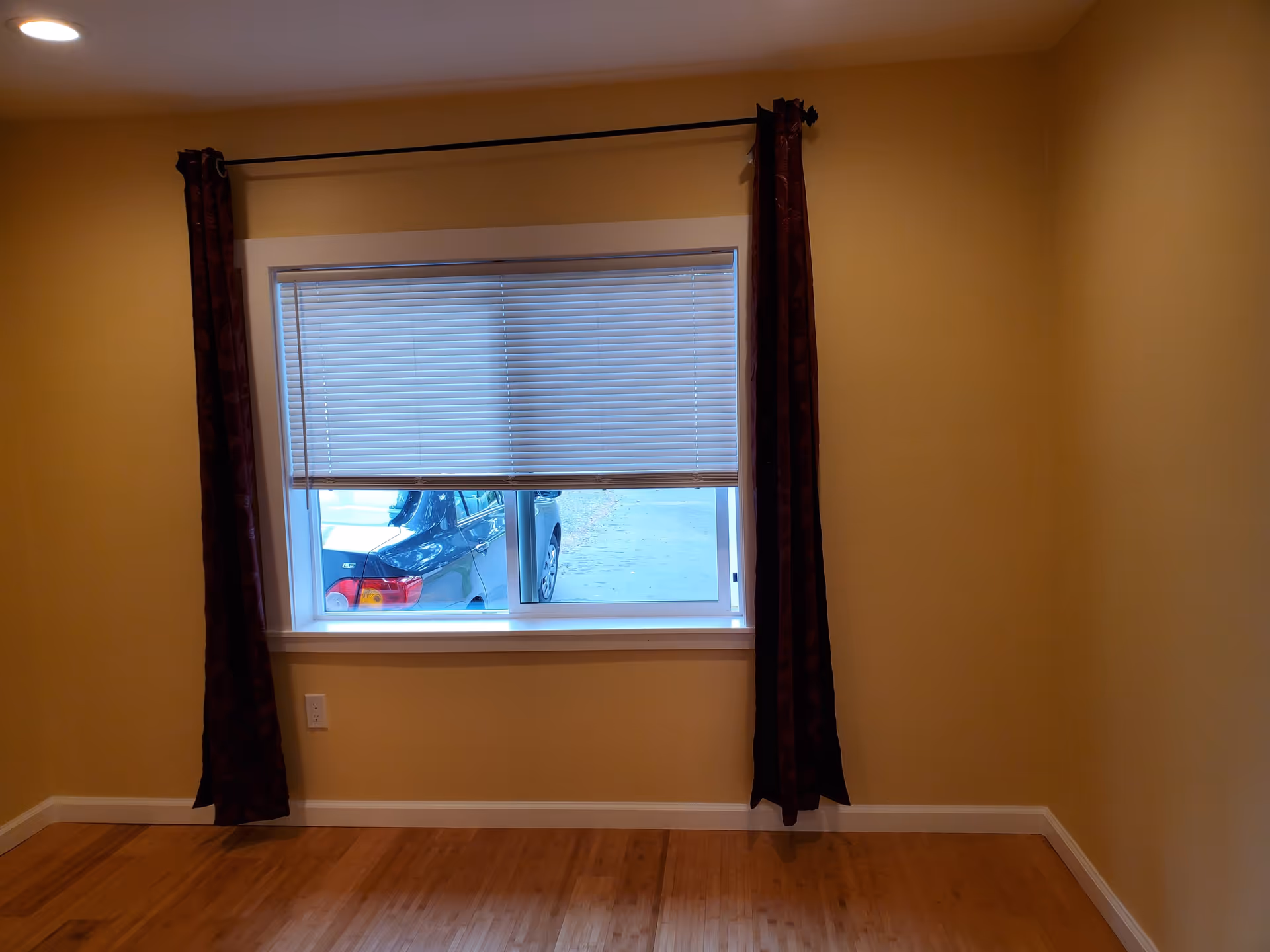 Empty room with light yellow walls, a wooden floor, and a window with white blinds partially open. Dark red curtains hang on either side of the window, and a black car is visible outside through the window.