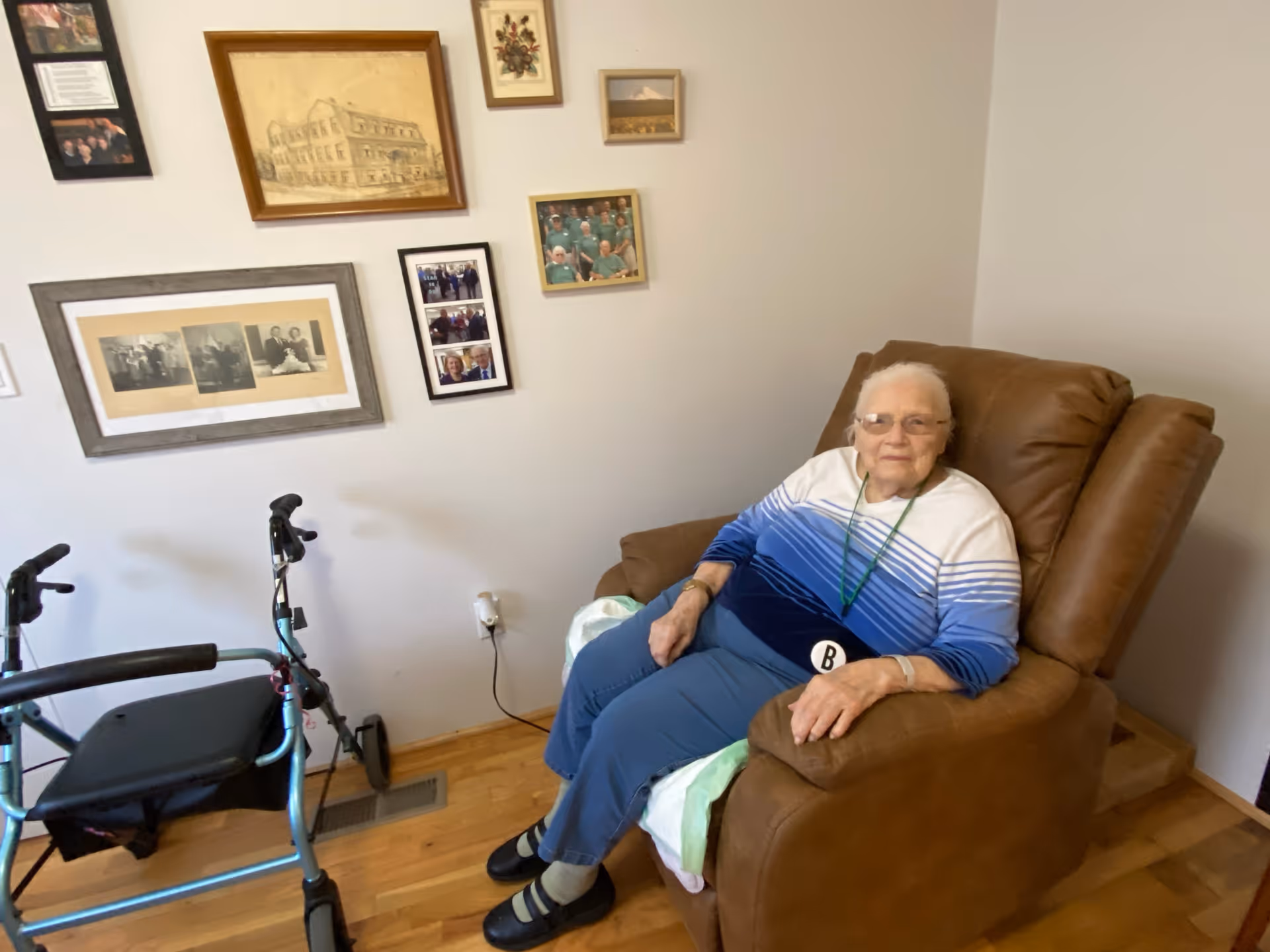 An elderly woman wearing glasses and a blue and white striped sweater sits comfortably in a brown recliner chair in a room with wooden flooring. Next to her is a blue walker. The wall behind her is decorated with several framed photographs and artwork.