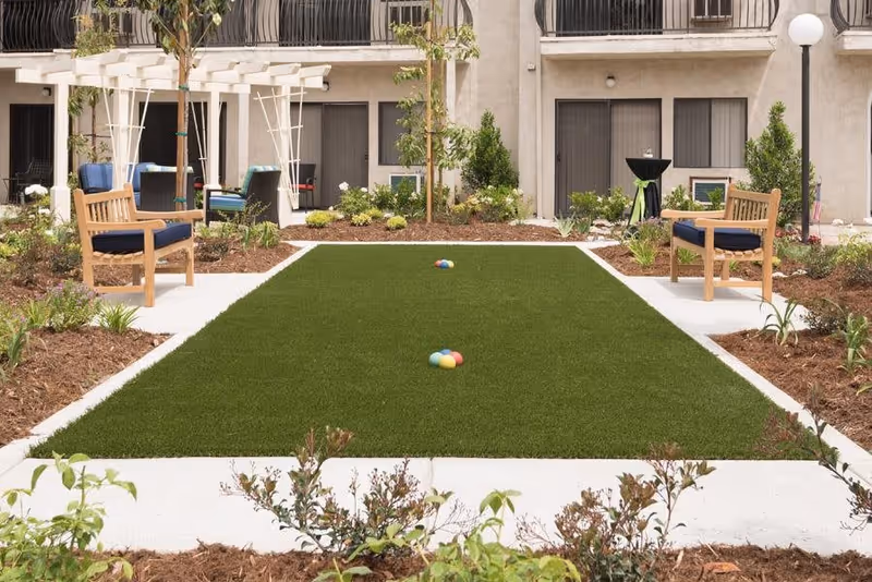 Outdoor garden area at Sunnycrest Senior Living with a rectangular green lawn bordered by concrete paths, two wooden benches with blue cushions on either side, small trees and plants surrounding the lawn, and a building with balconies and windows in the background.