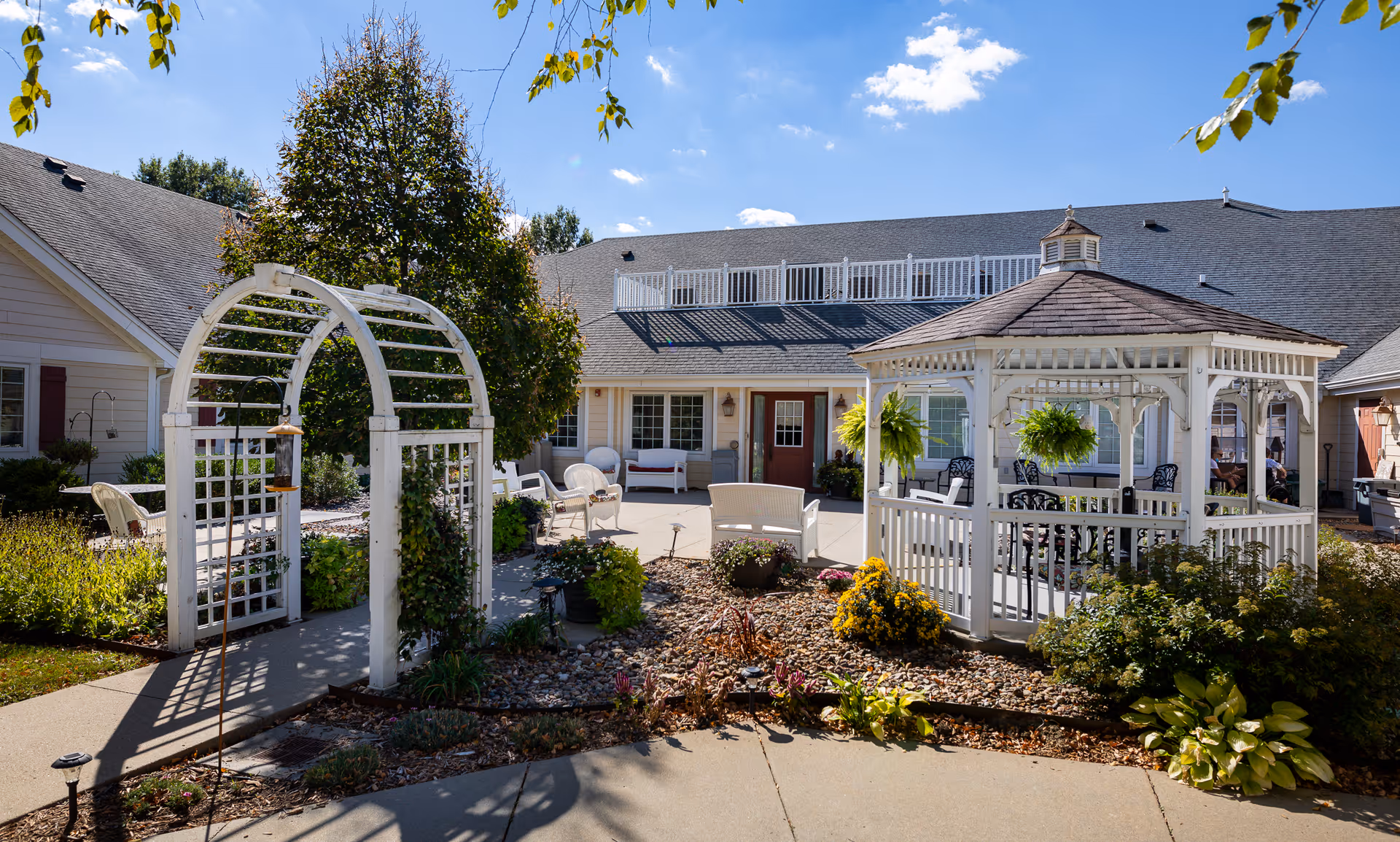 Sunny outdoor courtyard with a white gazebo, arched trellis, patio seating, and landscaped plantings in front of the facility building.