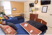Two hospital-style beds in a bright shared resident bedroom with a wall-mounted TV, dresser, lamps, and a window.
