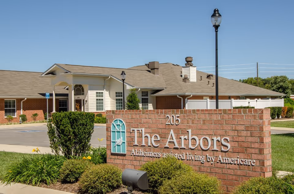 Exterior view of The Arbors, an Alzheimer's assisted living facility by Americare, showing a brick sign with the facility name and a single-story building with a covered entrance, surrounded by greenery and a clear blue sky.
