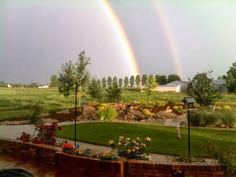 A landscaped backyard with lawn, flower beds, a pond-like rock feature and a double rainbow arching across the sky.