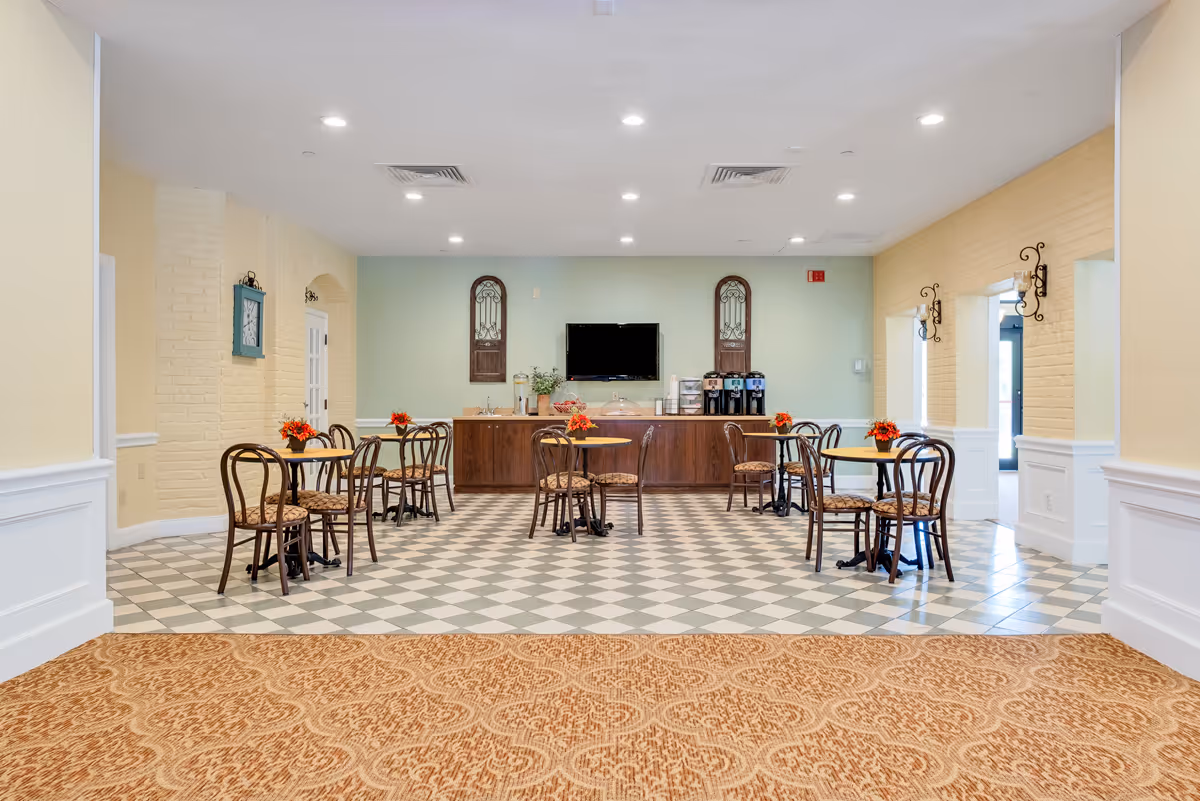 Bright communal dining area with small round tables and chairs facing a service counter with a TV and beverage station.